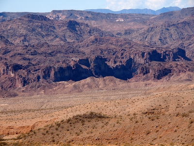 [Very dark brown rock formations with sandy colored ground dotted with small vegetation in the foreground.]